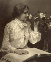 Helen Keller sitting with a large braille book open on her lap and she is smelling a rose that is being held in her left hand